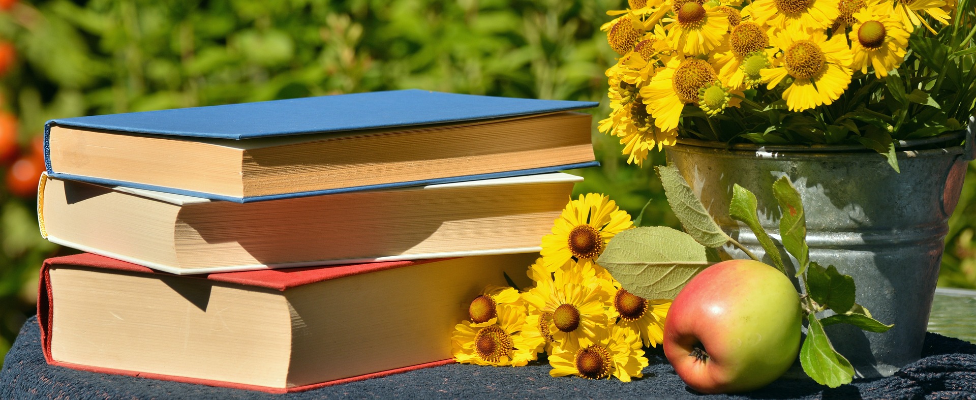 Foto von drei Büchern, gelben Blumen in einem Blecheimer und einem Apfel